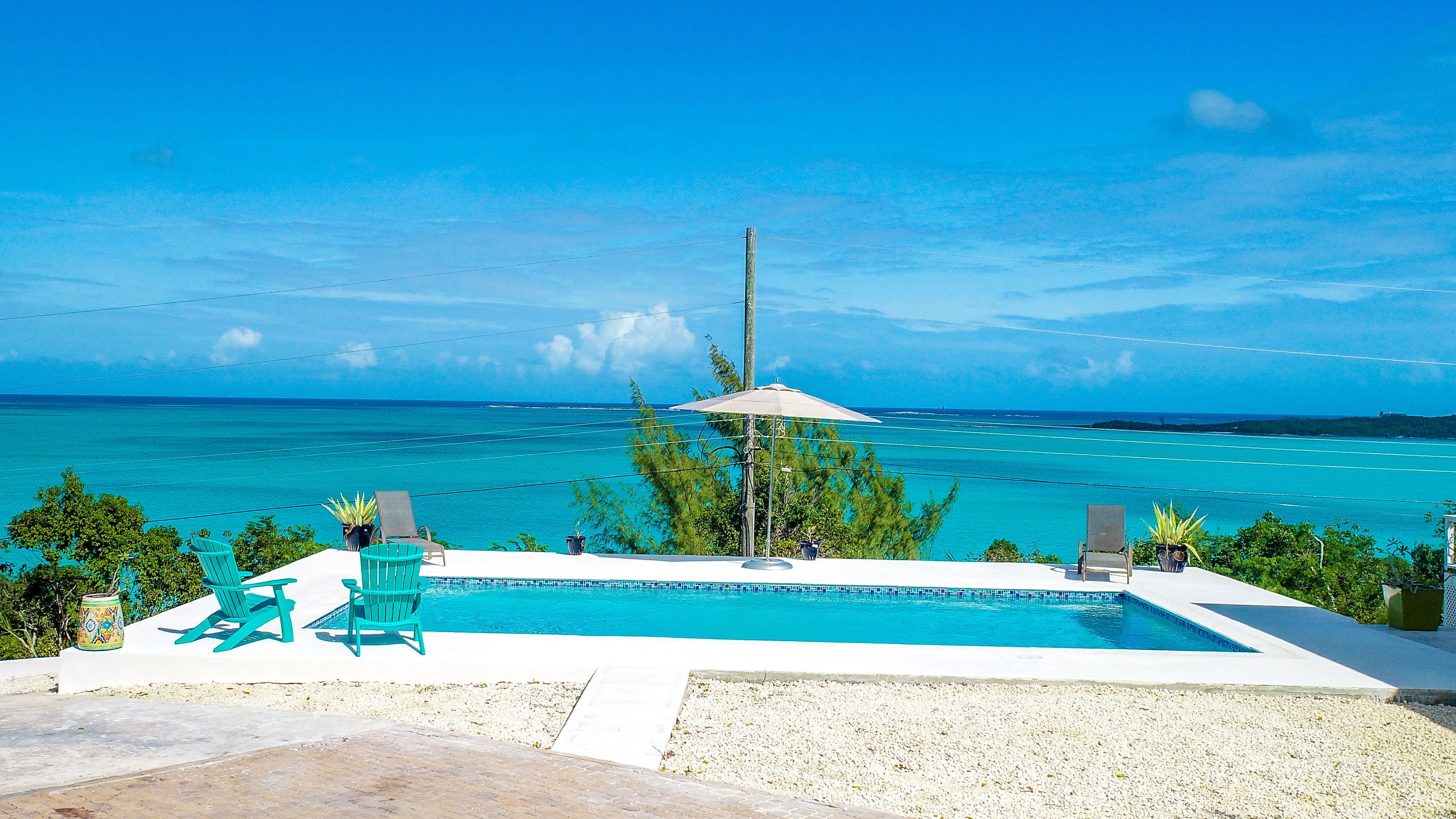 A pool with a view of the ocean, surrounded by chairs and a small umbrella.