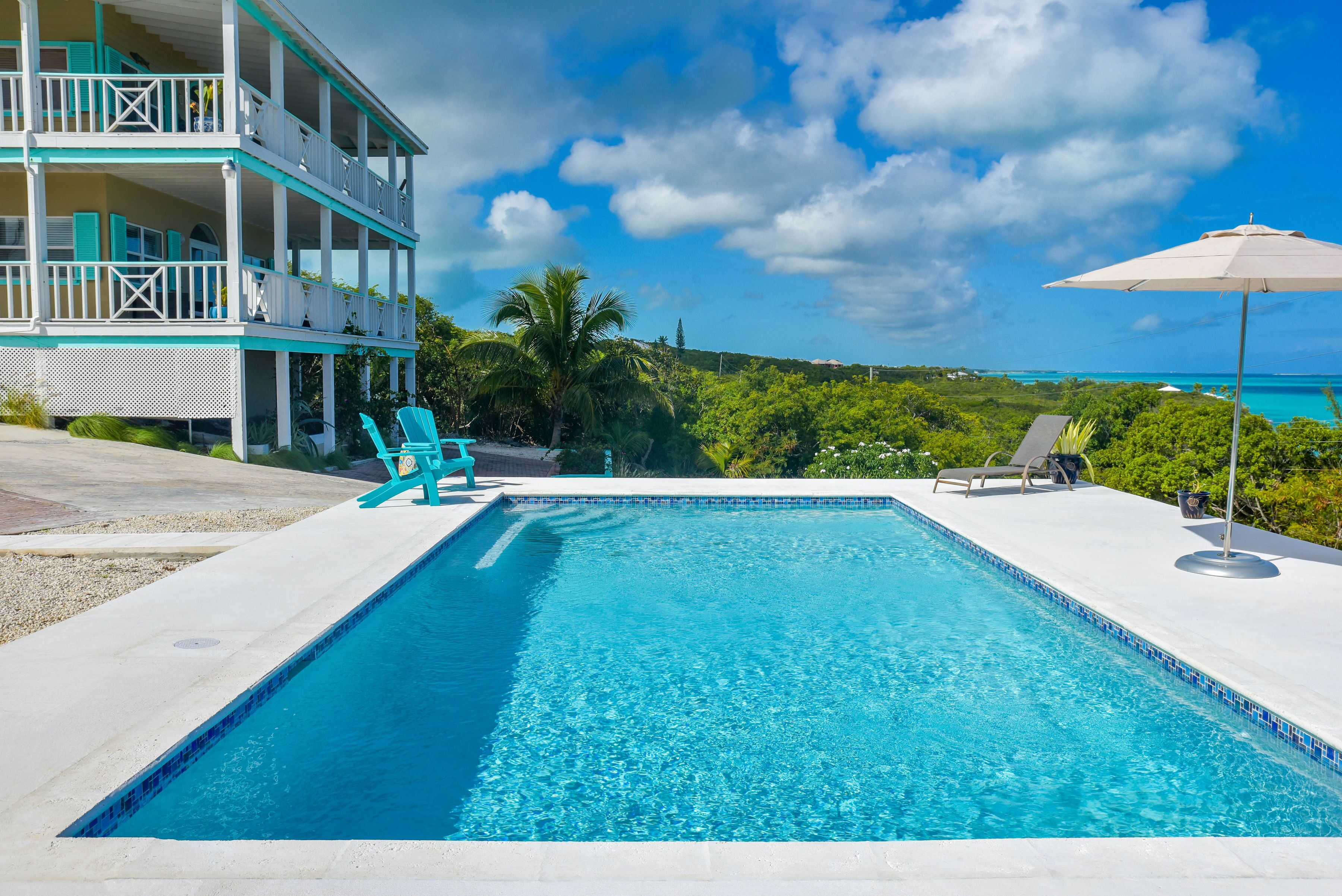 A swimming pool with a view of the ocean, a two-story building with balconies, and a beach chair.