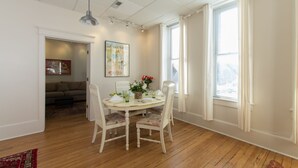 A dining area with a round table, white chairs, and a vase of flowers. There is a painting on the wall and a window with blinds.