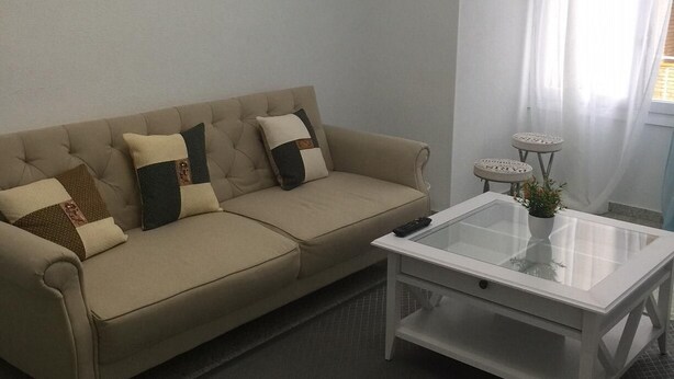 Una sala de estar con un sofá color beige, una mesita de centro de vidrio y una pequeña mesa auxiliar con una planta.