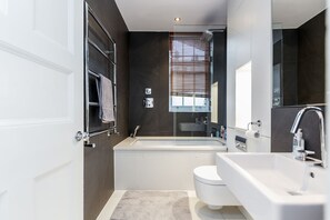 A modern bathroom with a bathtub, toilet, and sink. - Harrods Luxury Apartments (London)