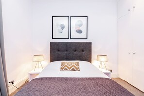 A neatly arranged bedroom with a bed, two bedside lamps, a grey upholstered headboard, and two framed abstract art pieces on the wall. - Harrods Luxury Apartments (London)
