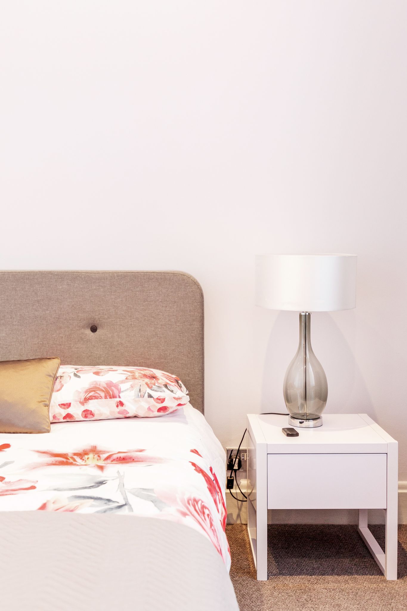 A neatly made bed with a floral bedspread and a single pillow, a white bedside table with a lamp, and a power outlet.