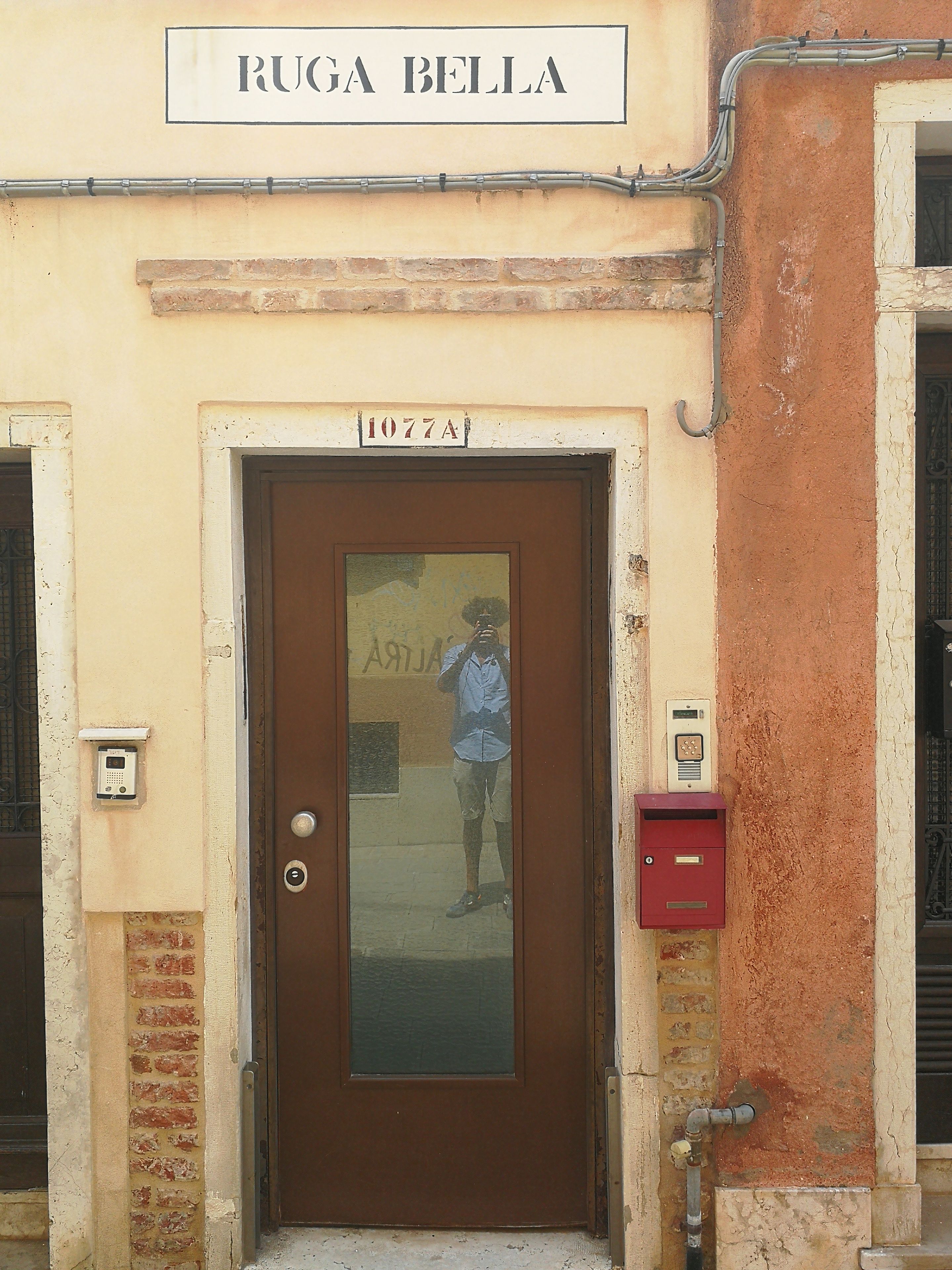 Entrada de un edificio con un letrero que dice "RUGA BELLA" y una puerta marcada con el número "1077A".