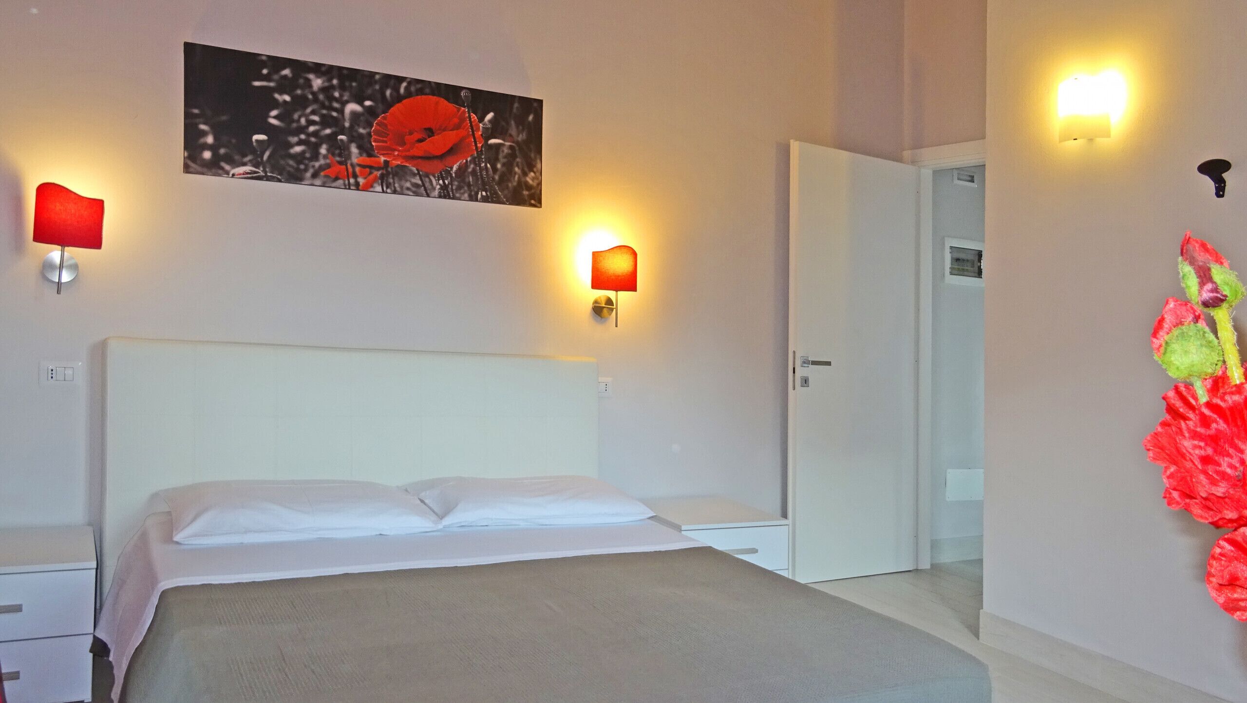 A hotel room with a bed, two bedside tables, a wall-mounted artwork of red flowers, and two wall sconces.