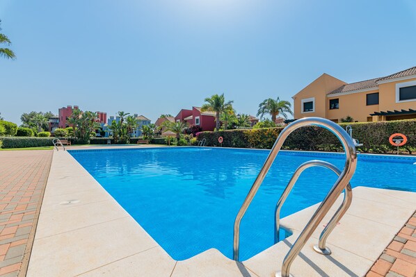Una piscina con una escalera de metal, rodeada por un camino de ladrillos y palmeras.