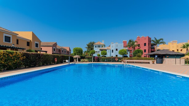 A clear blue swimming pool surrounded by colorful buildings with a clear sky. - El Rincon del Golf I - Isla Canela Golf GRHI6P06 (Isla Canela)