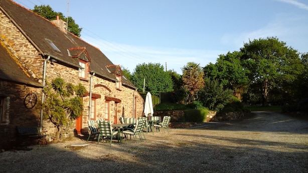 A stone building with a gravel driveway, outdoor seating area with chairs and a table, and a covered umbrella. - Lavender gite Le Cosquer St Connec (Saint-Connec)