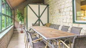 A covered patio with a wooden table and wicker chairs, stone walls, and a large window.