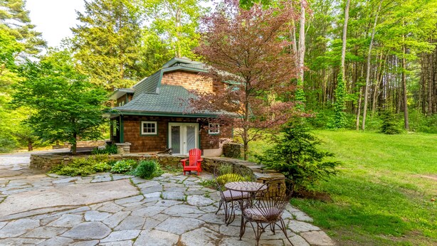 A stone patio with outdoor furniture, a small brick house with a green roof, and a forested area.