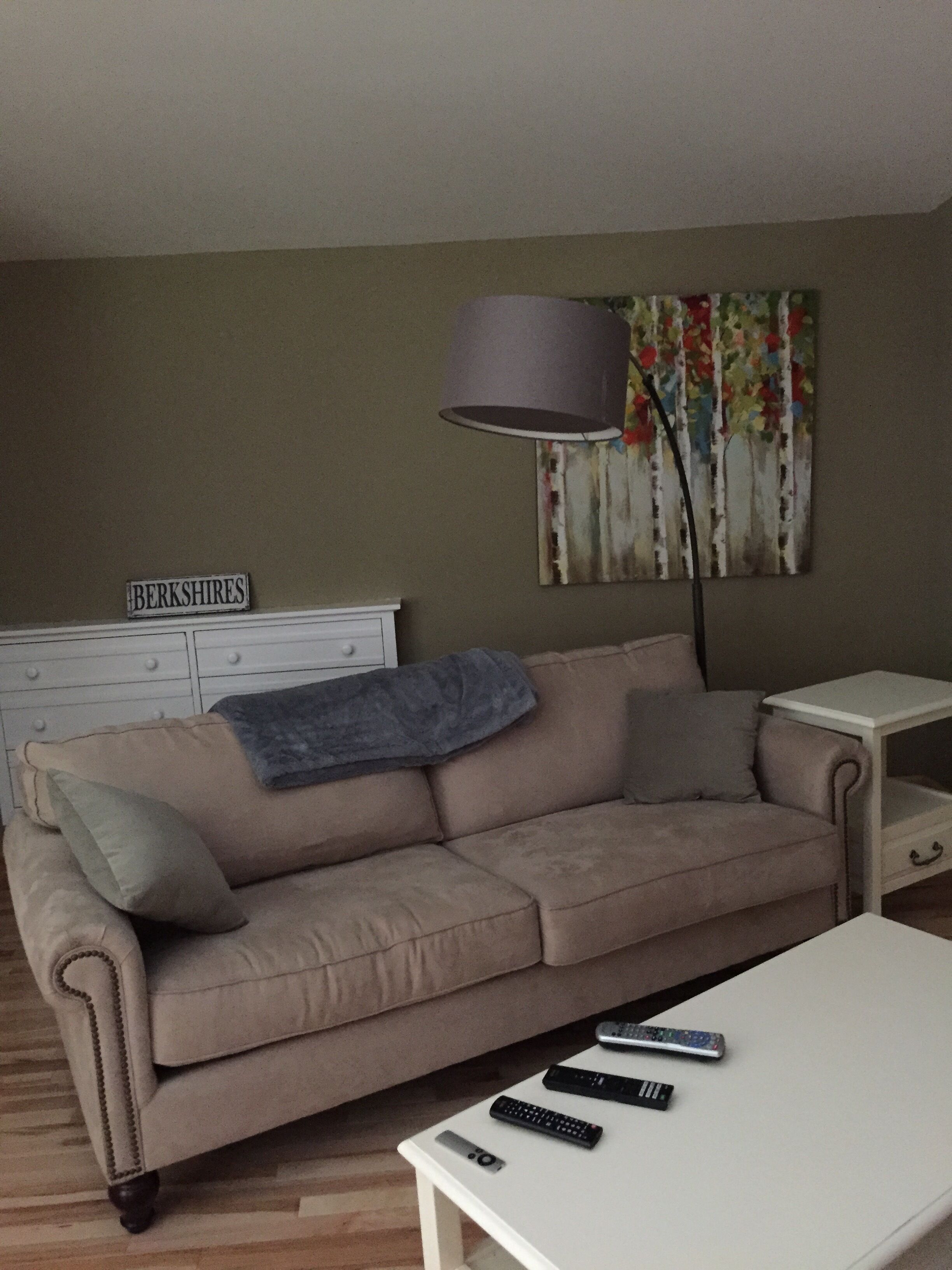 Ein Wohnzimmer mit einem beigen Sofa, einem weißen Couchtisch und einer Lampe. Auf dem Tisch liegen Fernbedienungen.