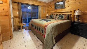 Une chambre à coucher située dans une cabane en bois, avec un lit, un ventilateur de plafond, une table de chevet, une chaise et une vue sur l’extérieur, visible à travers une porte vitrée.