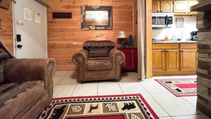Un salon chaleureux avec un fauteuil en cuir, un mur en bois, un petit meuble rouge et un coin cuisine doté de meubles en bois et d’un plan de travail.
