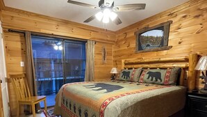 Une chambre à coucher dans une cabane en bois, comprenant un lit, une chaise, une table de chevet, un ventilateur de plafond et une fenêtre avec des rideaux.