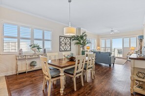 A spacious dining area with a wooden table and chairs, a large window with blinds, and a ceiling fan. - Oceanfront Corner Condo with Private Balcony Pool Hot Tub and Garage Parking (New Smyrna Beach)