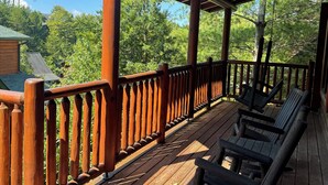 Une terrasse en bois avec une rambarde, des chaises en bois et une vue sur des arbres et un bâtiment en arrière-plan.