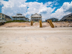 A beach house with a central structure, two staircases leading to a sandy beach, and surrounding buildings.