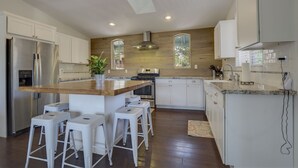 Une cuisine moderne avec des meubles blancs, un îlot central en bois, des tabourets de bar, des appareils électroménagers en acier inoxydable et une grande fenêtre qui laisse entrer la lumière naturelle.