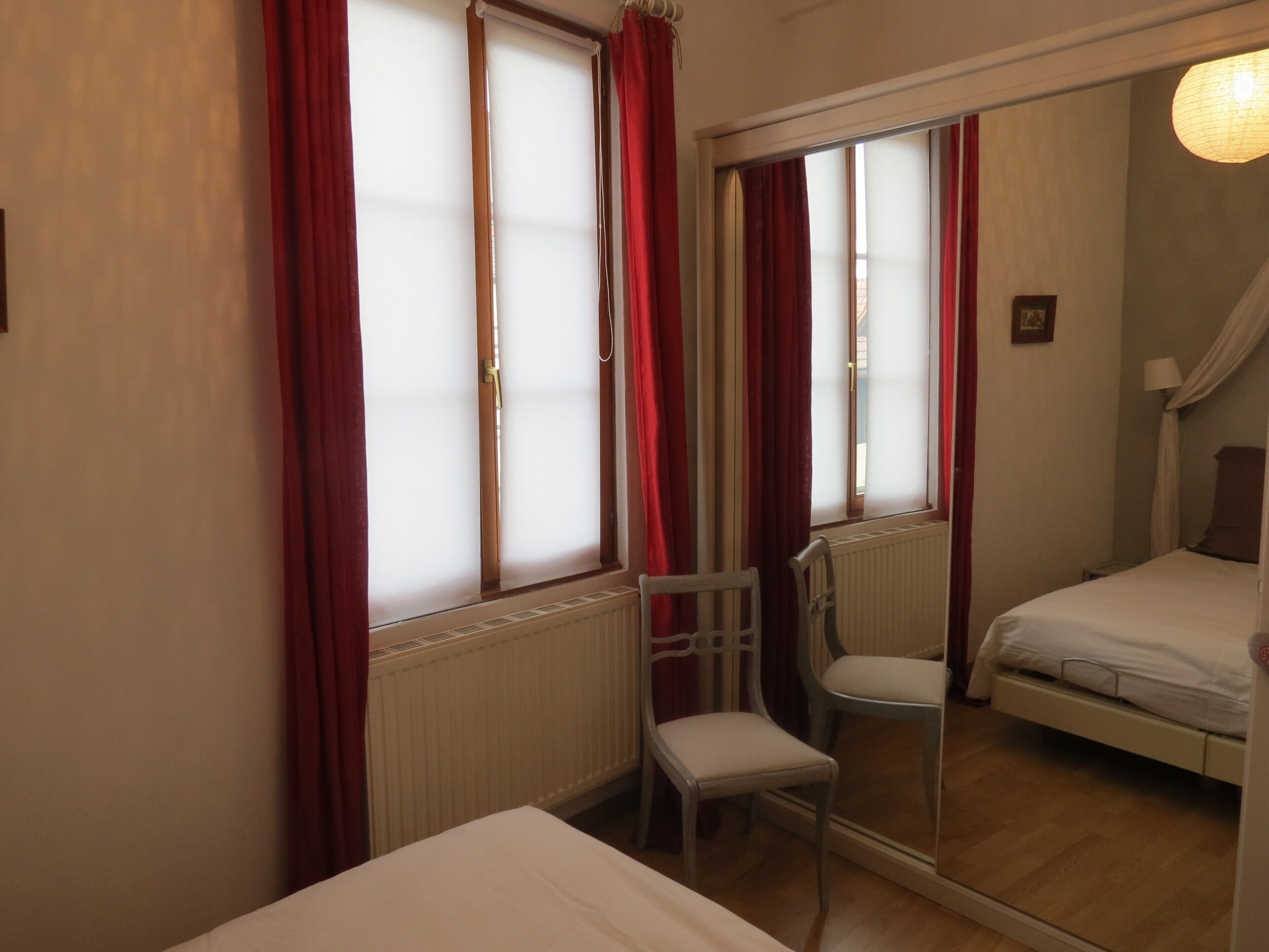 A bedroom with a bed, a chair, a window with curtains, and a mirror.