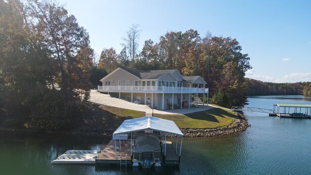 A large house with a covered dock and a boat, surrounded by trees and a lake.