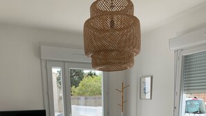 Ein Zimmer mit Holztisch, Fernseher auf einem Schrank, einem Korbstuhl und einem großen Fenster mit Jalousien.