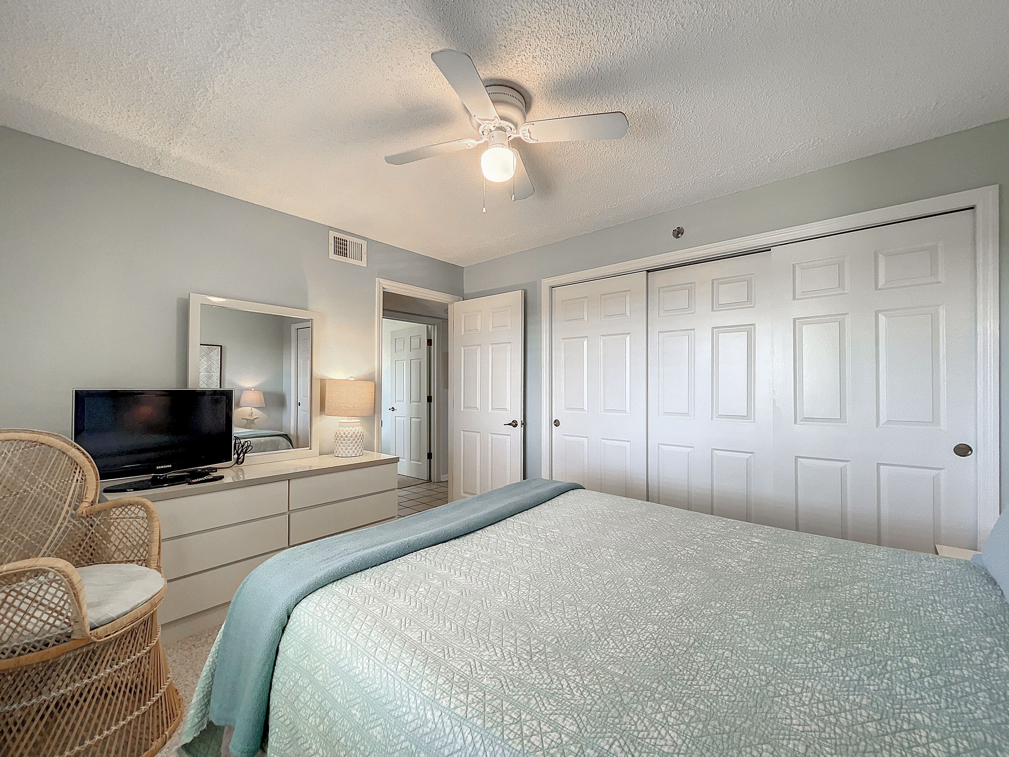 A bedroom with a bed, a television, a ceiling fan, a door, and a chair.