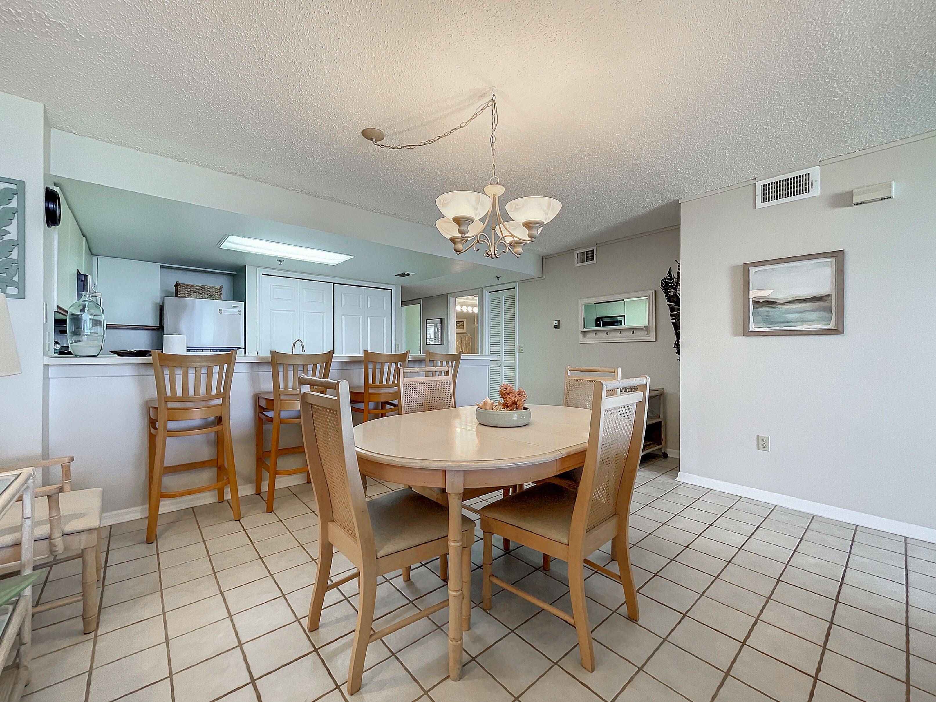 A dining area with a round table and six wooden chairs, a chandelier, and a kitchen in the background.