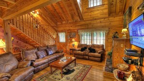 A cozy log cabin interior with a wooden staircase, leather sofas, a television, and a fireplace. - Bears Den, Pigeon Forge, Arcade game, Hot Tub (Pigeon Forge)