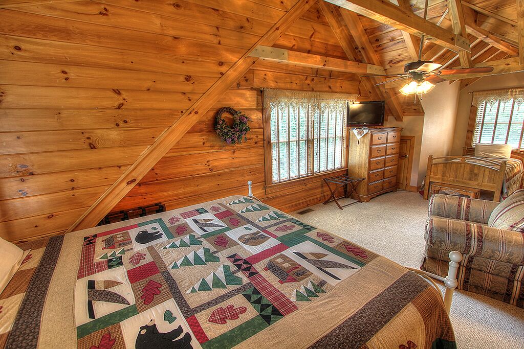 A cozy log cabin bedroom with a large bed, a dresser, a television, and a ceiling fan.
