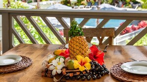 A wooden table with a pineapple centerpiece, surrounded by tropical fruits and flowers, set on a balcony with a view of a pool and greenery. - 2026 Waikoloa Hilton pool pass included 6 guests daily, available through 2026! (Waikoloa Village)