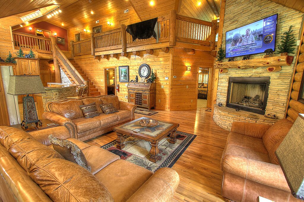 A log cabin interior with a wooden staircase, leather sofas, a fireplace, and a television mounted on the wall.