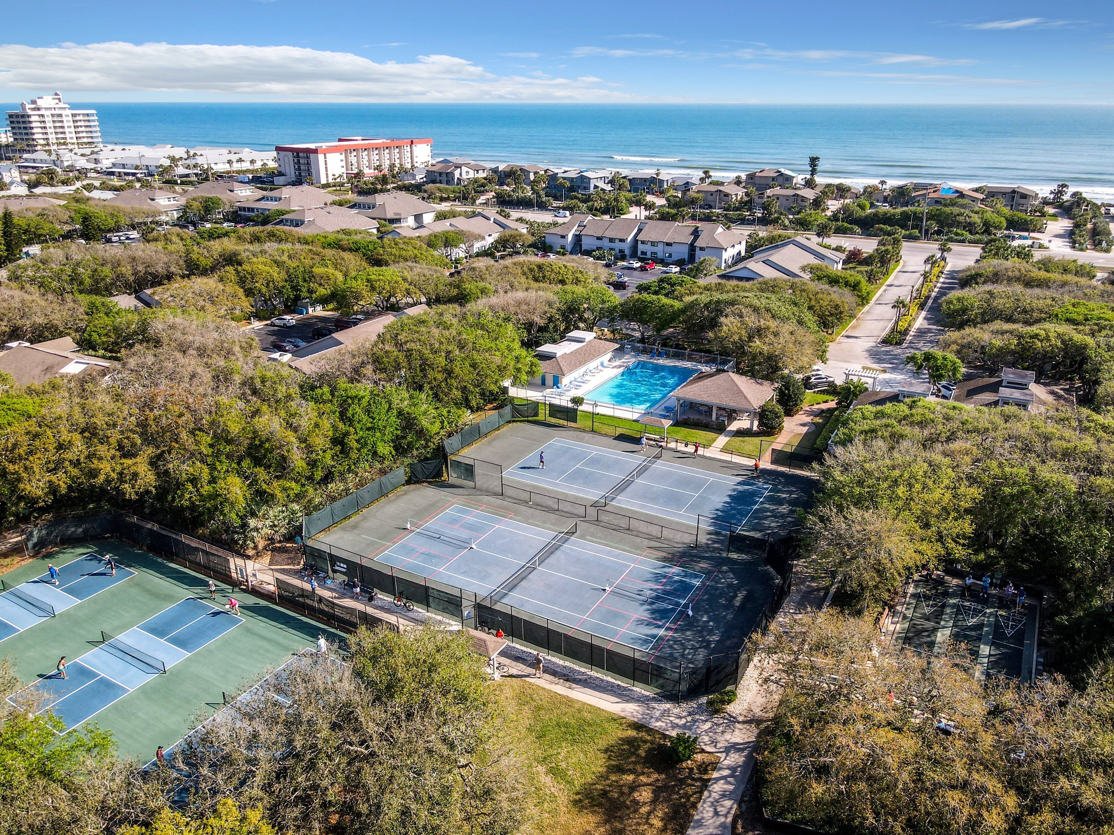 An aerial view of a residential area with a large swimming pool, tennis courts, and a view of the ocean.
