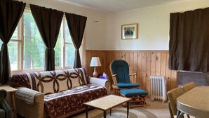 Ein gemütliches Wohnzimmer mit Sofa, Sessel und Tisch, Holzwänden und einem fenster mit Vorhängen.