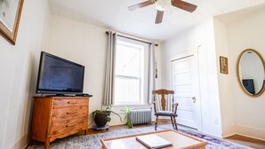 Una habitación con televisión sobre un mueble de madera, una silla, una mesita de café, un ventanal con cortinas, un radiador y un ventilador de techo.
