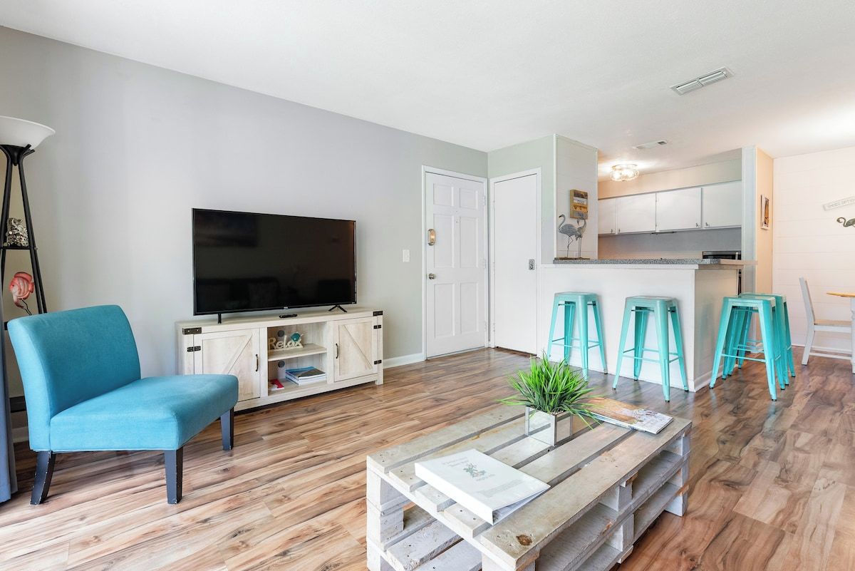 A living room with a television, a sofa, a coffee table, and a kitchen area with stools.