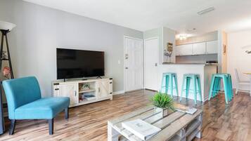 A living room with a television, a sofa, a coffee table, and a kitchen area with stools.
