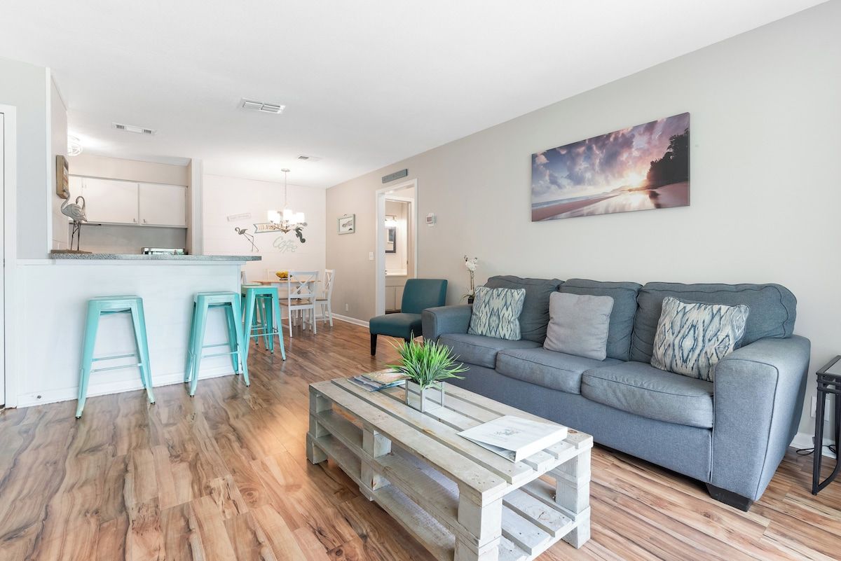 A modern living room with a grey sofa, teal bar stools, a wooden coffee table, and a painting of a sunset on the wall.