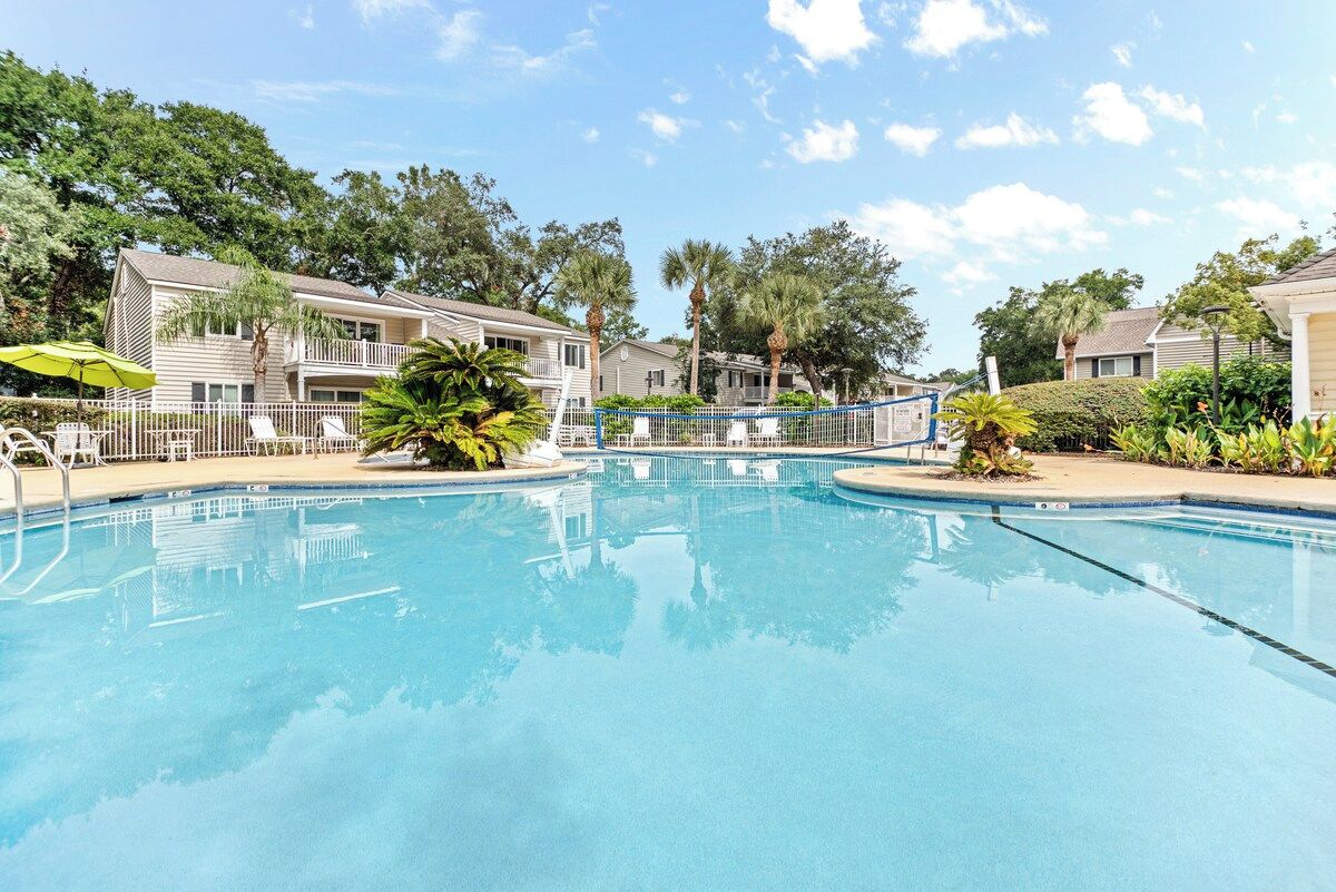 A swimming pool surrounded by buildings with balconies, trees, and a clear sky.