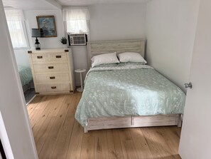 A bedroom with a wooden bed frame, a bedside table, a dresser, and a window with a curtain. - The Cabin on Chappy! (Edgartown)