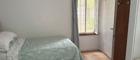 A room with a bed, a window with curtains, a wooden floor, and a door.
