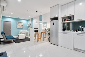 A modern kitchen with white cabinets, a microwave, and a refrigerator. There is a dining area with a table and chairs, and a living area with a sofa and a plant.