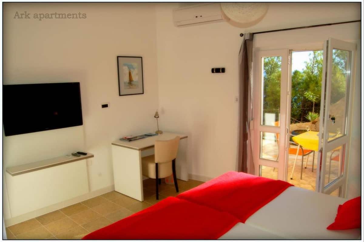 A bedroom with a bed, a desk, a chair, a TV, and a sliding glass door leading to an outdoor area.