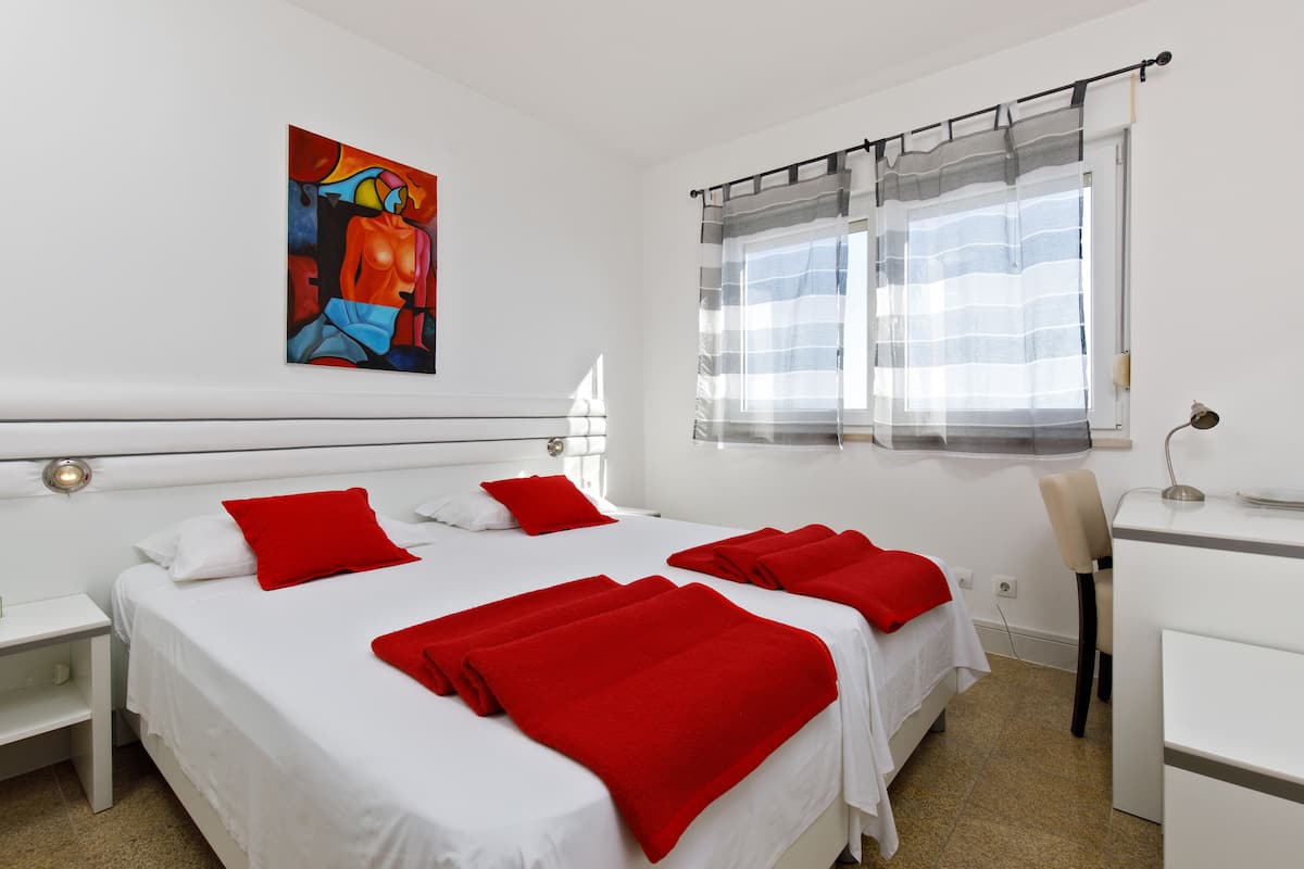 A hotel room with a large bed, two red pillows, a white headboard, a painting on the wall, a window with curtains, and a desk with a chair.