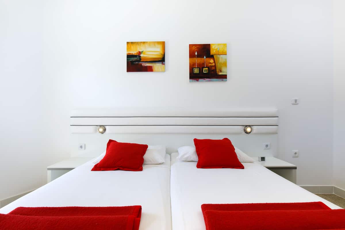 A hotel room with two beds, white bedding, red pillows, and two paintings on the wall.