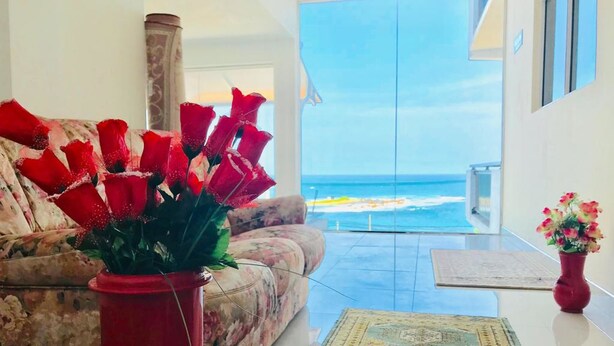 A living room with a sofa and a red vase of flowers, a view of the ocean, and a balcony with a glass door. - Queens Beach Hotel (Tangalle)