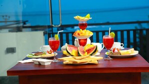 A table set with breakfast items including toast, fruit, and drinks, with a view of the sea in the background. - Queens Beach Hotel (Tangalle)