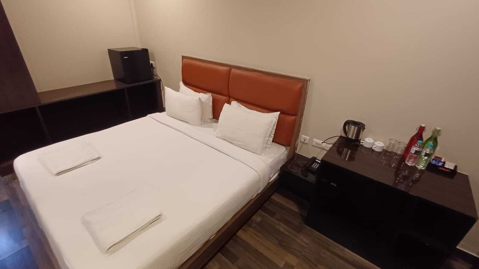 A hotel room with a bed, a nightstand, a coffee maker, and a small table with bottles and glasses.