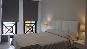 Ein modernes Schlafzimmer mit einem großen Bett, zwei Nachttischlampen, einem kleinen Nachttisch und Blick durch ein Fenster mit geometrischem Muster.