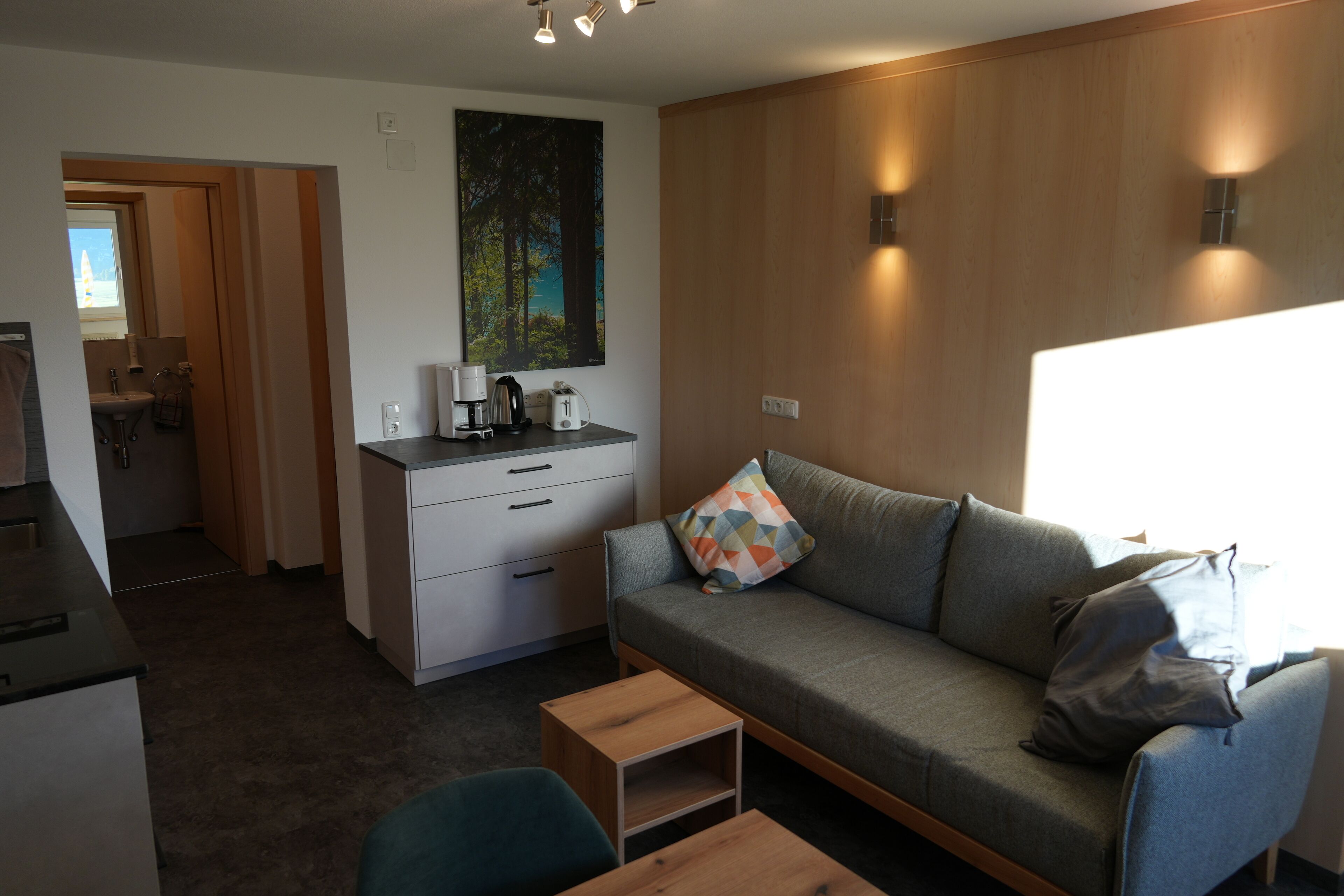 A modern living room with a grey sofa, a small wooden table, and a kitchen area in the background.