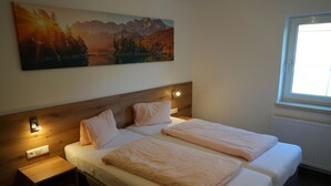 A bedroom with two beds, a nightstand, a wall-mounted light, and a painting of a mountain landscape. - Ferienhaus Mott (Lermoos)